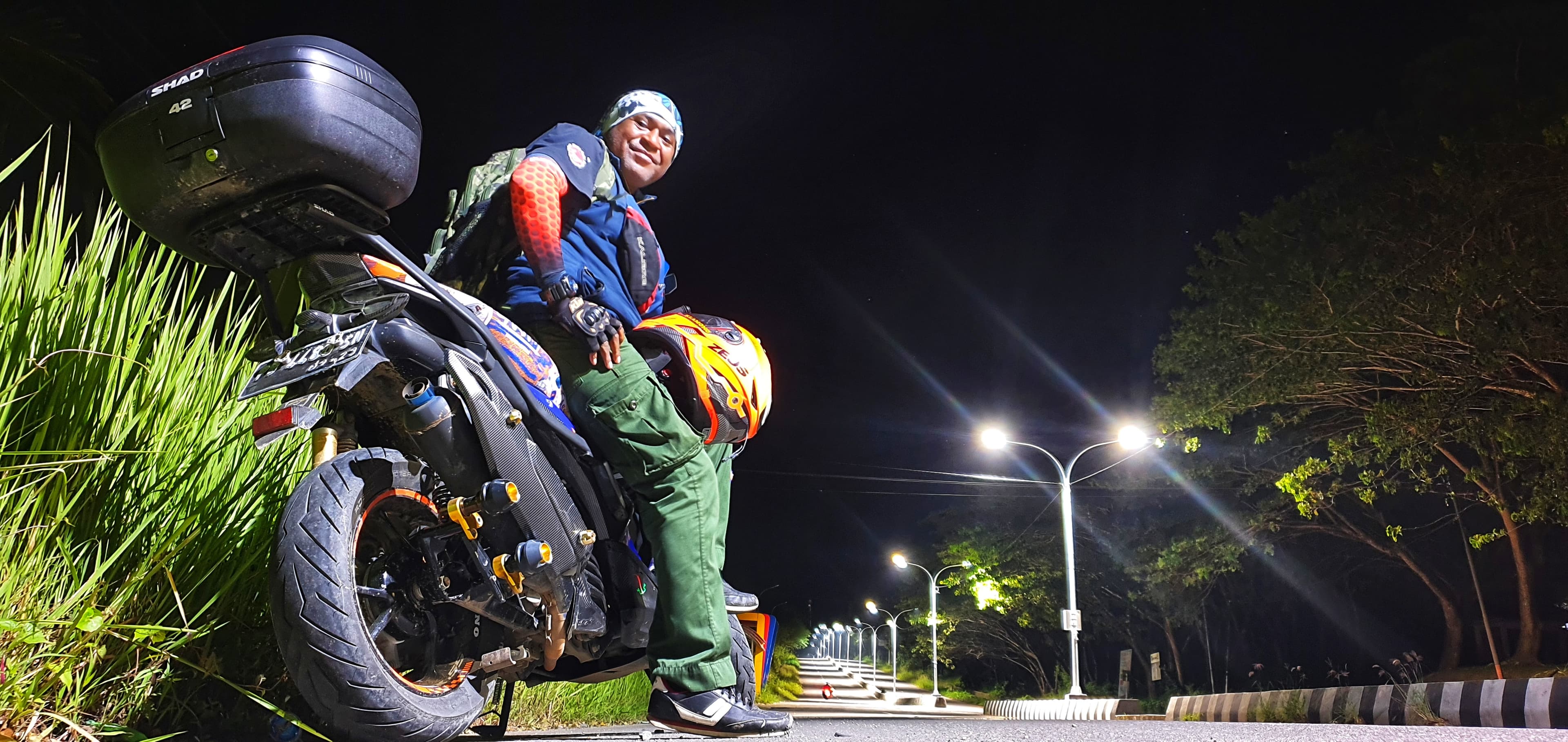 A man sits on a motorcycle at night, holding an orange helmet under streetlights.