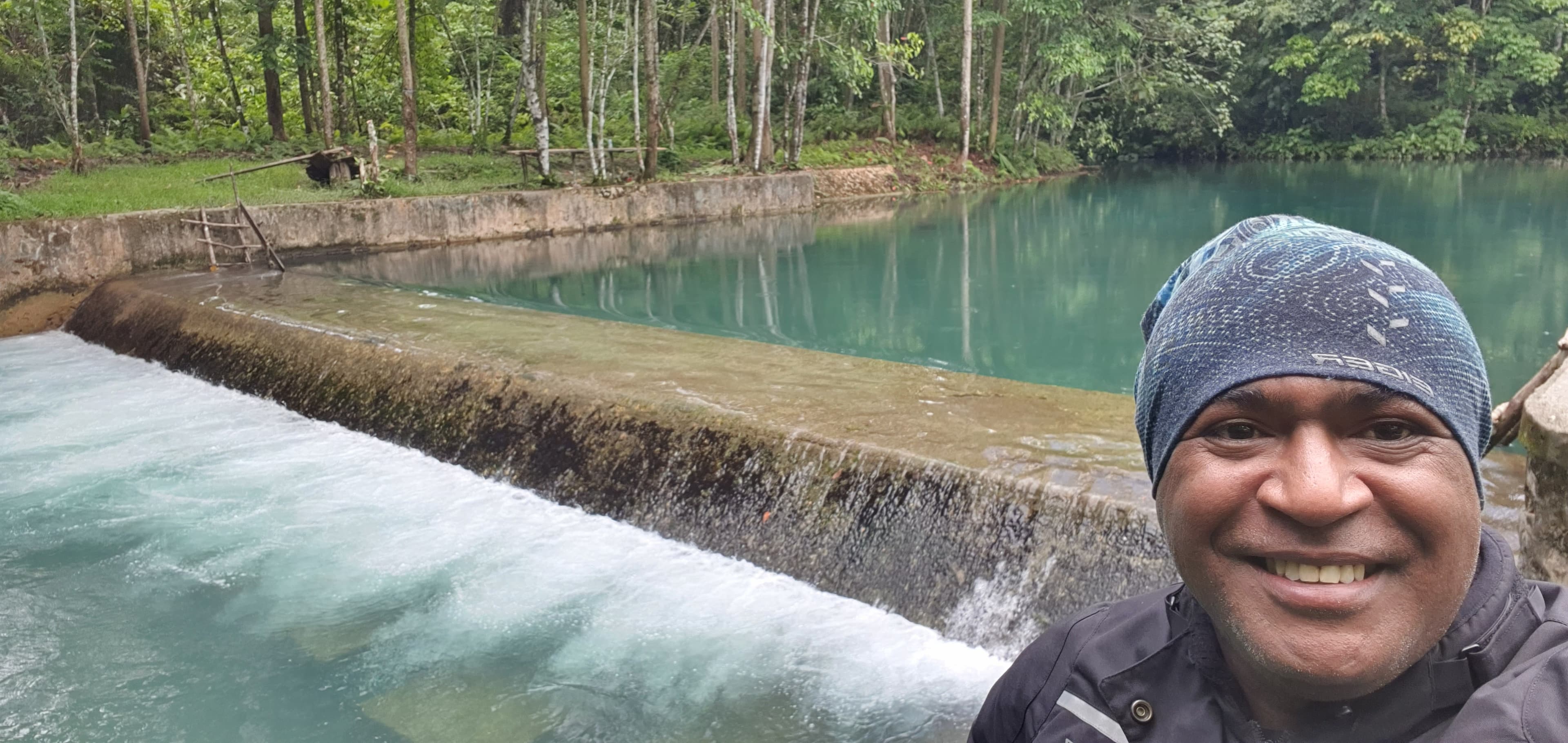 Smiling man in a blue beanie before a turquoise forest pool and small concrete dam.