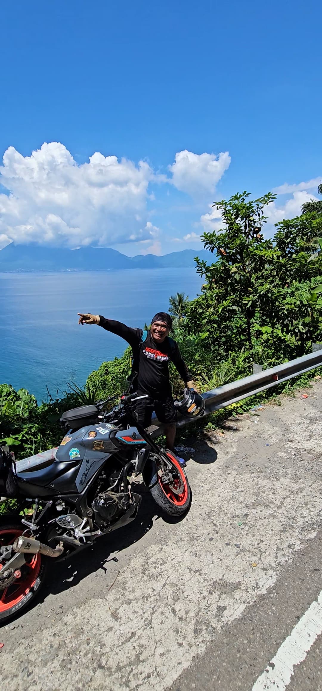 Smiling man points at a scenic ocean view while standing beside his parked motorcycle.