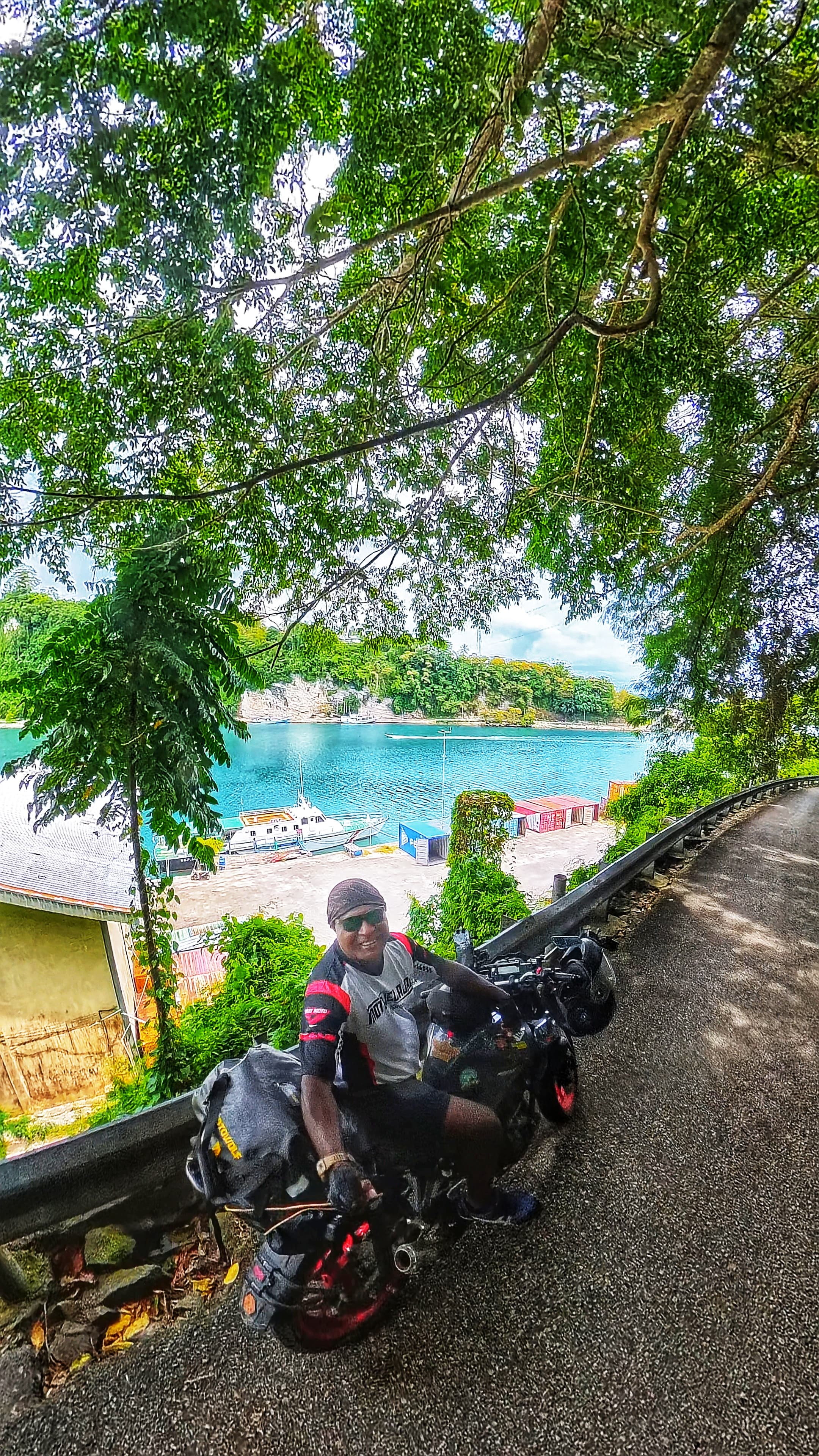 Motorcyclist on a scenic road overlooking a turquoise bay with boats and lush greenery.